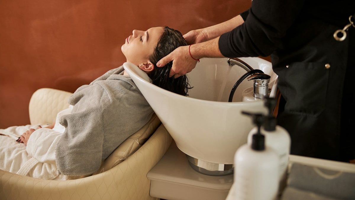 Kundin mit geschlossenen Augen am Waschbecken im Friseursalon, die Friseurin massiert ihr die Kopfhaut beim Haarewaschen
